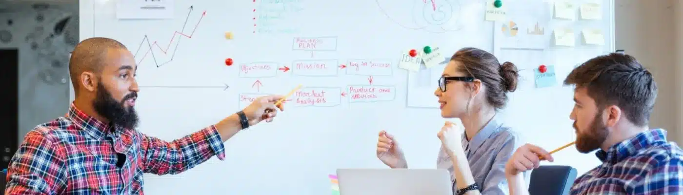 Image of a group of people around a whiteboard. On the left is a man pointing, on the right is a man and woman listening.