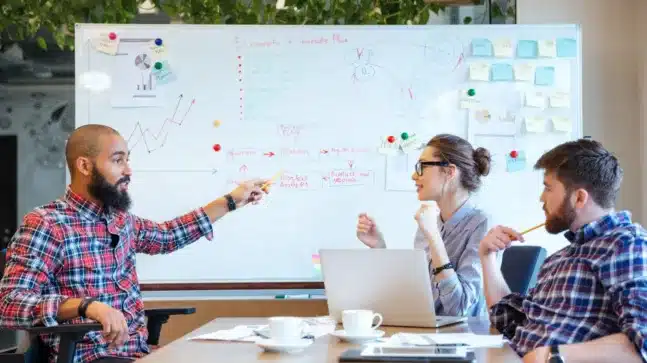 Image of a group of people around a whiteboard. On the left is a man pointing, on the right is a man and woman listening.